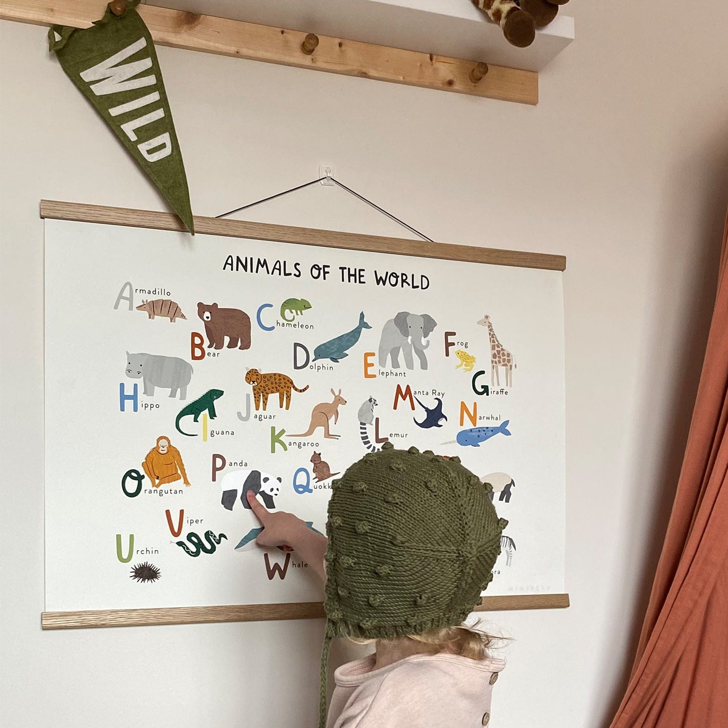 Art Print Animals Of The World Alphabet Art Print on a wall being pointed at by a toddler with a WILD customisable Arnold and the Caterpillar pennant in green above