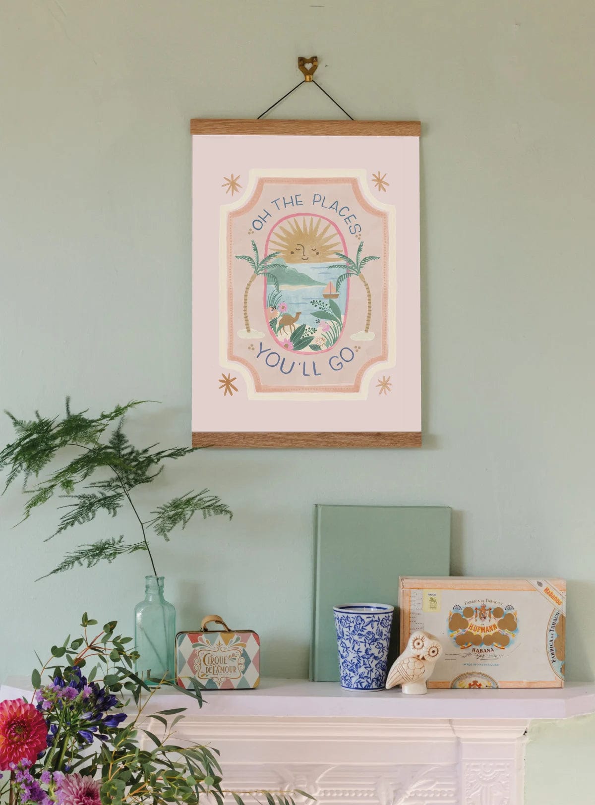 The top of a vintage iron fire place, with a white mantle, with boho style accessories on , including a miniature suitcase and a book. Hanging from a picture hook in an oak hanger is our oh the places you'll go art print. giving you are my sunshine vibes. 