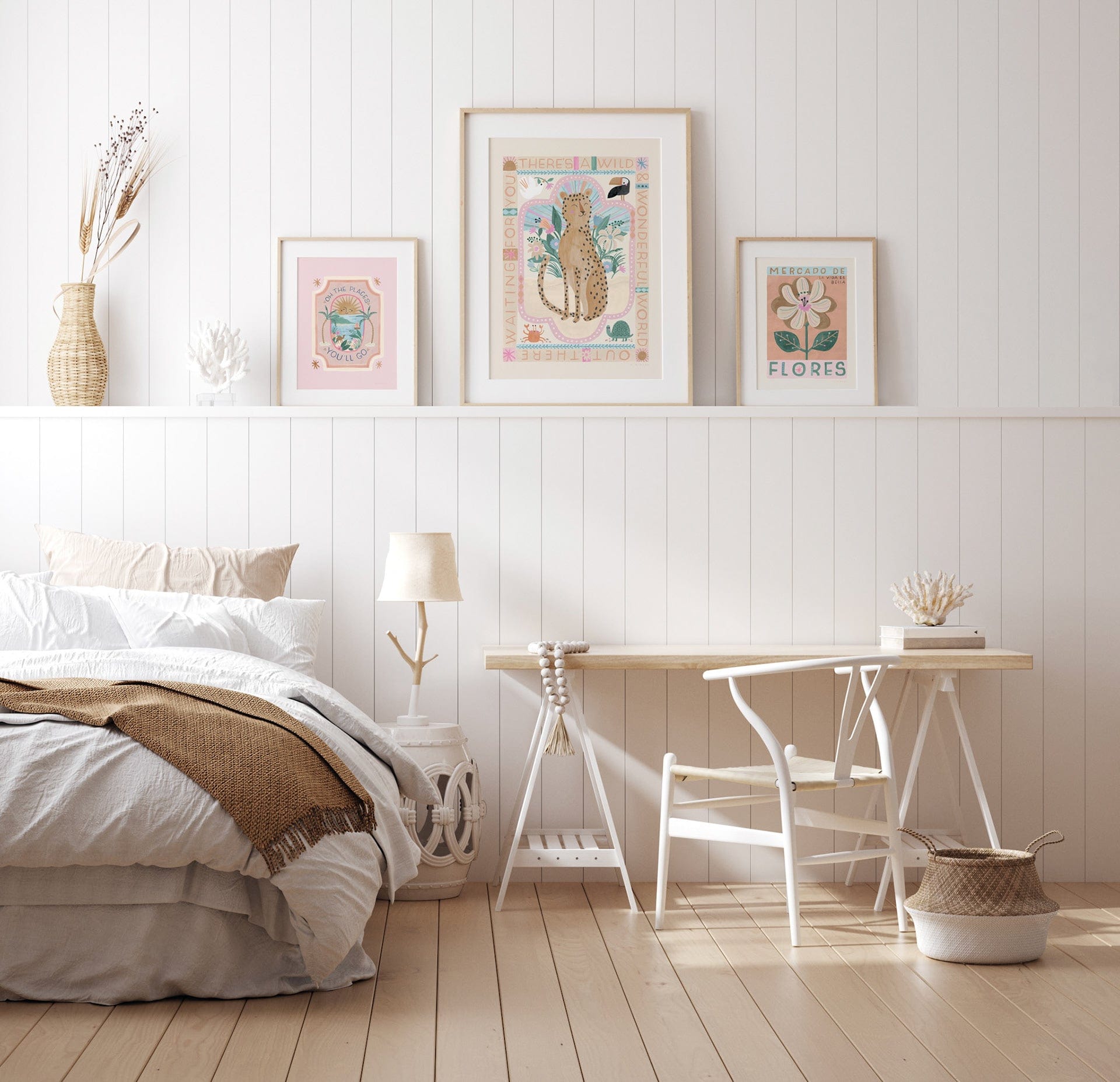 A scandi style bedroom showing a comfy bed with a white barrel night stand, and a white and light wood trestle table desk with a white chair and basket on the floor. On the shelf behind the bed are 3 art prints in light wood frames, the large print in the centre being our wild, wonderful, world leopard art print, and either side are smaller art prints, our oh, the places you go and mercado de flores.