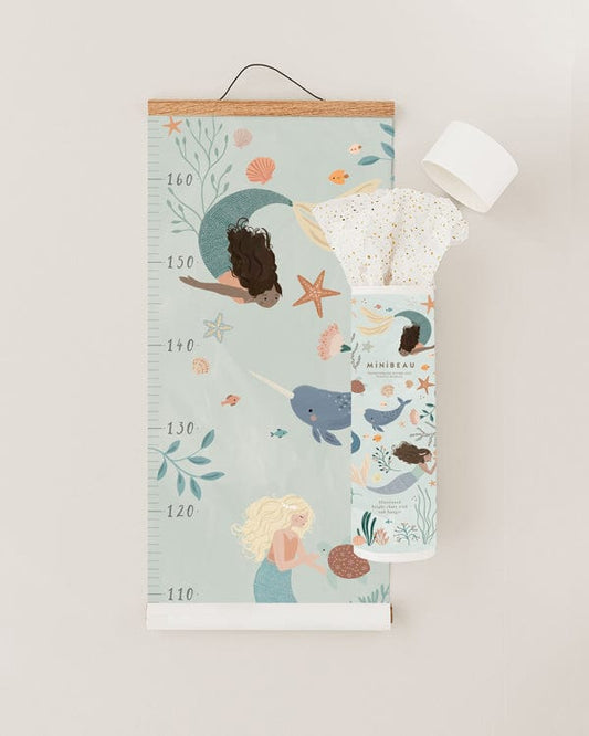 Photo showing our Mermaid height chart rolled up from the bottom to about half way with the open packaging tube overlapping it to the right. Growth chart features 2 mermaids, a narwhal, starfish, fish and sea foliage. The packaging white tube is printed with similar artwork and had gold speckled tissue paper coming out of the top.