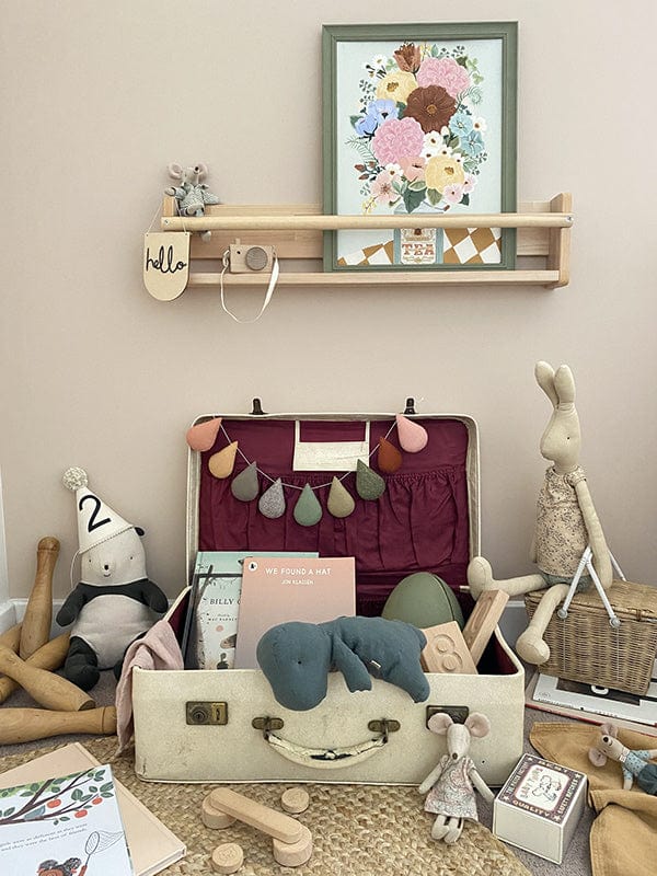 A vintage suitcase open, against a wall with toys and books spilling out of it, Maileg mice, a Maileg bunny, a soft panda with a party hat on, below a spice rack shelf with a maileg mouse, a hello wooden pennant, a wooden toy camera and our Boho Floral vase art print in a green frame. 