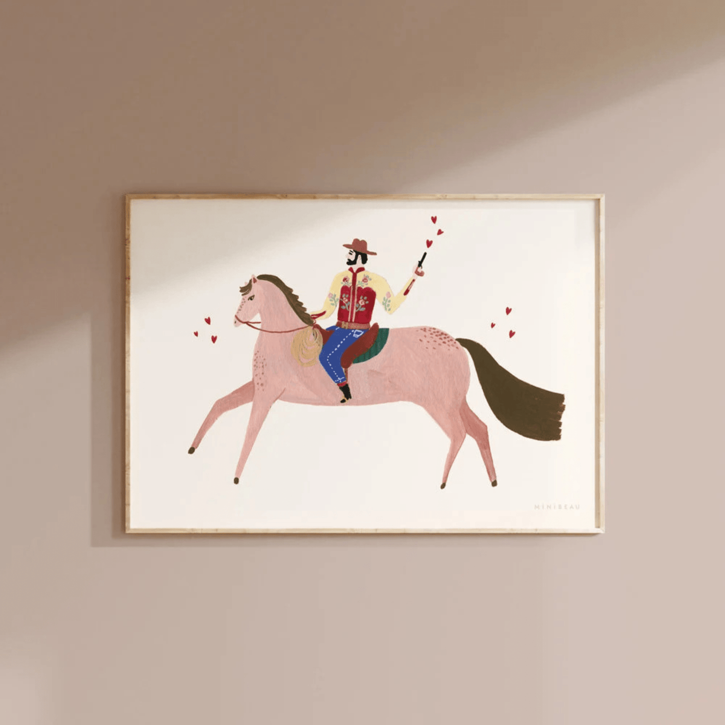 Art print hung on a light brown wall in a light wood frame. Our cowboy Art print features a cowboy, dressed in a floral shirt, on a brown horse, shooting red hearts from a pistol on a neutral background.