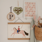 A row of white wooden hooks with a basket with a bunny in and our cowboy art print (in an oak hanger) hanging from it. Resting on the hooks is our Kristina art print, mounted in white board. An embroidered floral P and a green heart with birds in hangs on the wall.
