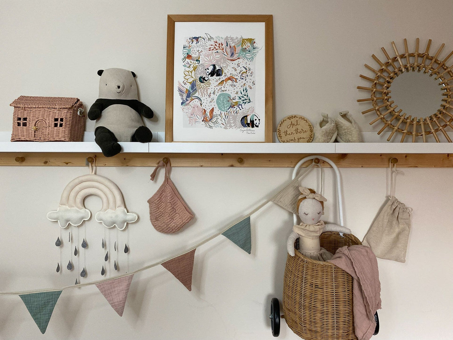 A wooden and white painted shelf in a kids space with hooks on the underside. With bunting, a rainbow mobile, a pink bonnet and a Olli Ella luggy with a doll, all in muted tones, underneath. On the shelf sit an Olli Ella Casa Clutch, a soft panda, a framed Crayon Wilderness art print, booties and a rattan framed mirror.