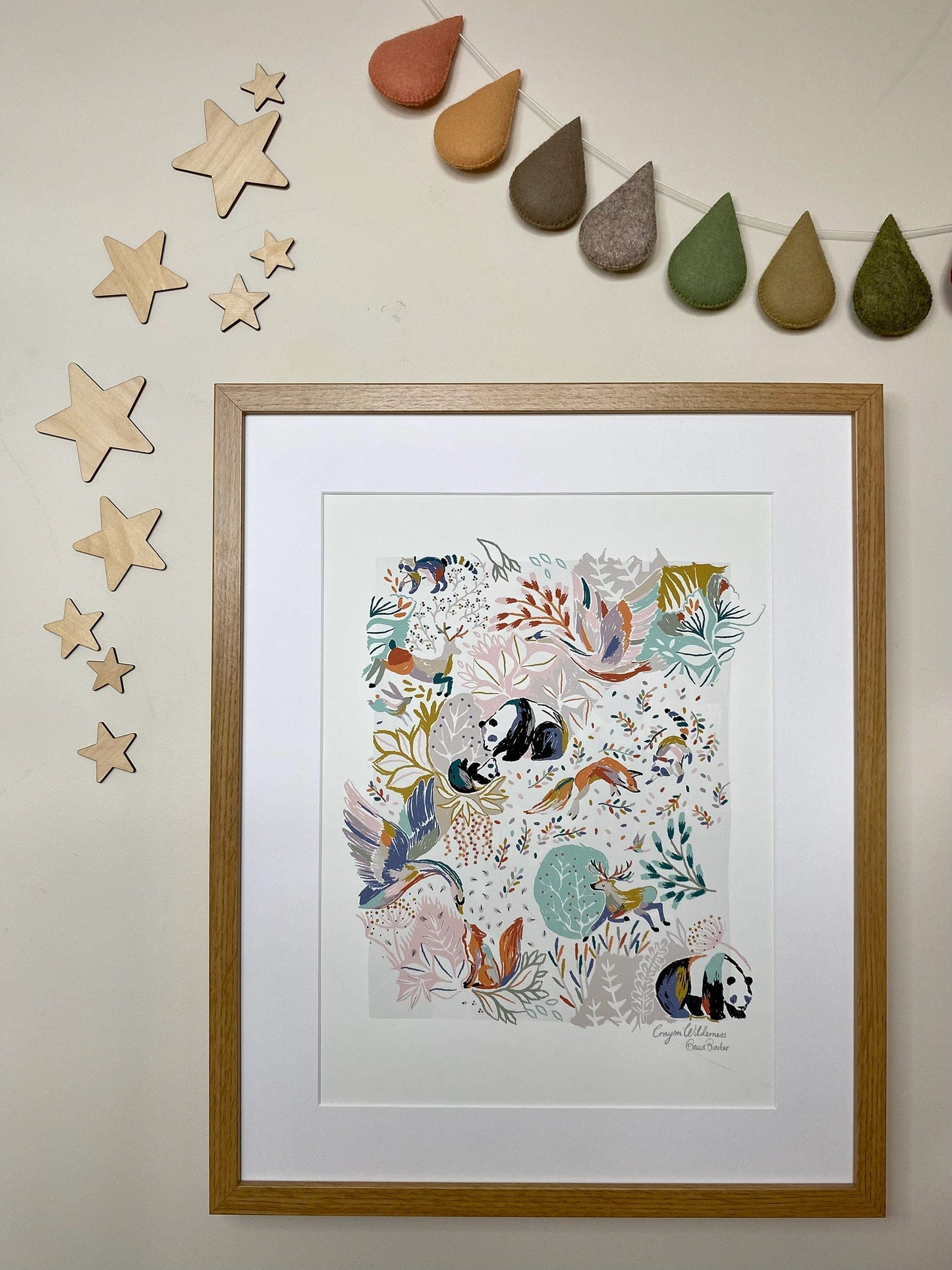 Our Crayon Wilderness art print in a dark oak frame hung on a neutral wall, with rain drop bunting in autumnal hues, and wooden stars framing the hung art work. Our Crayon Wilderness Art print is hand-drawn with floral details in greens and pinks, with pop of brighter colours, with animals featuring pandas, swans, foxes, deer and raccoons, creating a whimsical woodland feel.