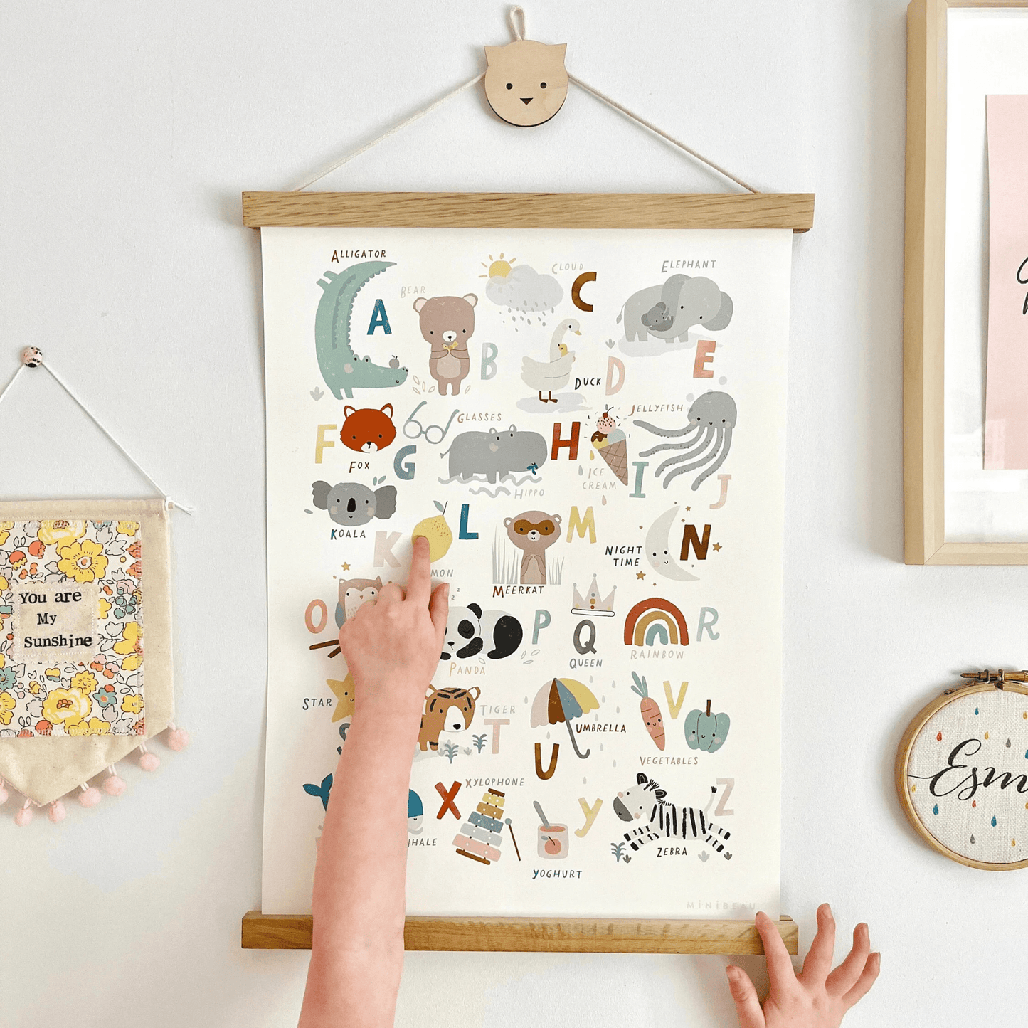 Image of a child pointing at a Lemon on our cute animals art print. Art print is hung in an oak hanger from a cat shaped wooden hook. With a 'you are my sunshine' pennant hanging next to it. Our Cute animals Alphabet Art print features a variety cute animals and everyday items in soft tones, representing every letter of the alphabet. The print includes everything from a rainbow and elephant to a star and panda, all set against a clean white background.