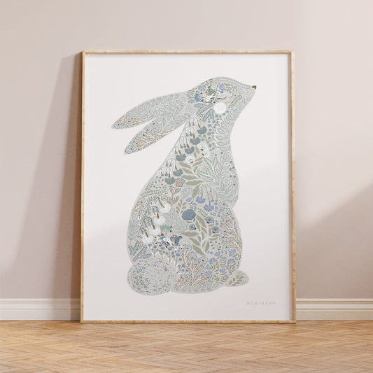 Photo showing our Floral Bunny Art Print in Blue in a light wood frame leaning against a neutral wall on a parquet flooring. Our Floral Bunny Art Print in Blue features a cute bunny sitting up with its back facing out so we can see it's tail, and it's head slightly turned towards the front so it's eye and nose can be seen. The Bunny has been drawn with ornate blooms and foliage filling it in, in blues and greens, on a neutral background.