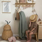 A child sitting on a wooden stool hiding their face with a straw hat, next to a ferm living pear basket and below a wooden shelf with hooks. An Olli Ella luggy hangs from the hooks with a doll and blanket hanging out of it, a wire-wrapped 'hello' sign, and bunting. On the shelf are wooden blocks, an Olli Ella Casa Clutch, a wooden flag saying autumn, and our Mercado de Flores art print in a frame. Hanging from an oak hanger from a round decorative hook is our floral bunny art print in blue. 
