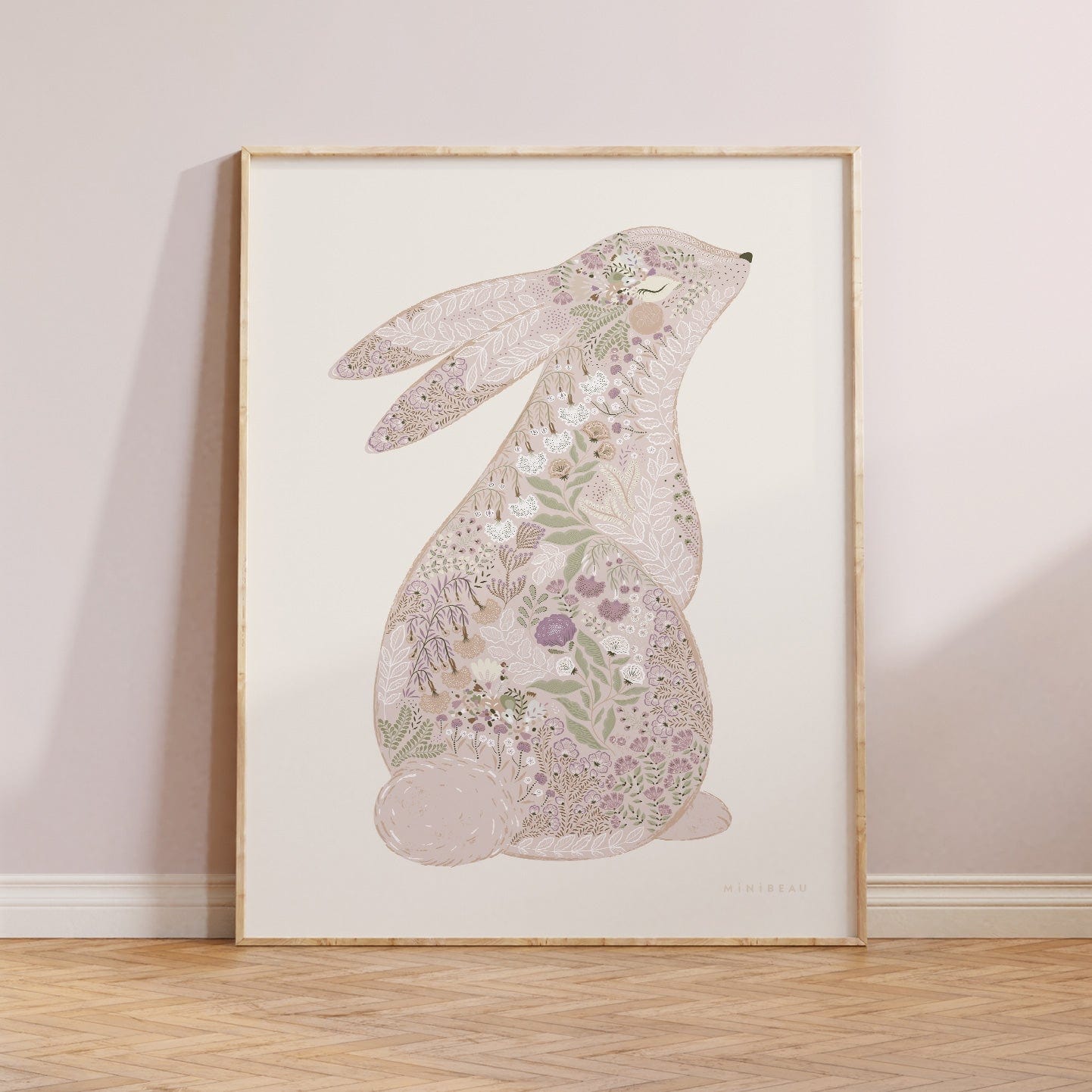 Photo showing our Floral Bunny Art Print in Pink in a light wood frame leaning against a neutral wall on a parquet flooring. Our Floral Bunny Art Print in Pink features a cute bunny sitting up with its back facing out so we can see it's tail, and it's head slightly turned towards the front so it's eye and nose can be seen. The Bunny has been drawn with ornate blooms and foliage filling it in, in Pinks and greens, on a neutral background.