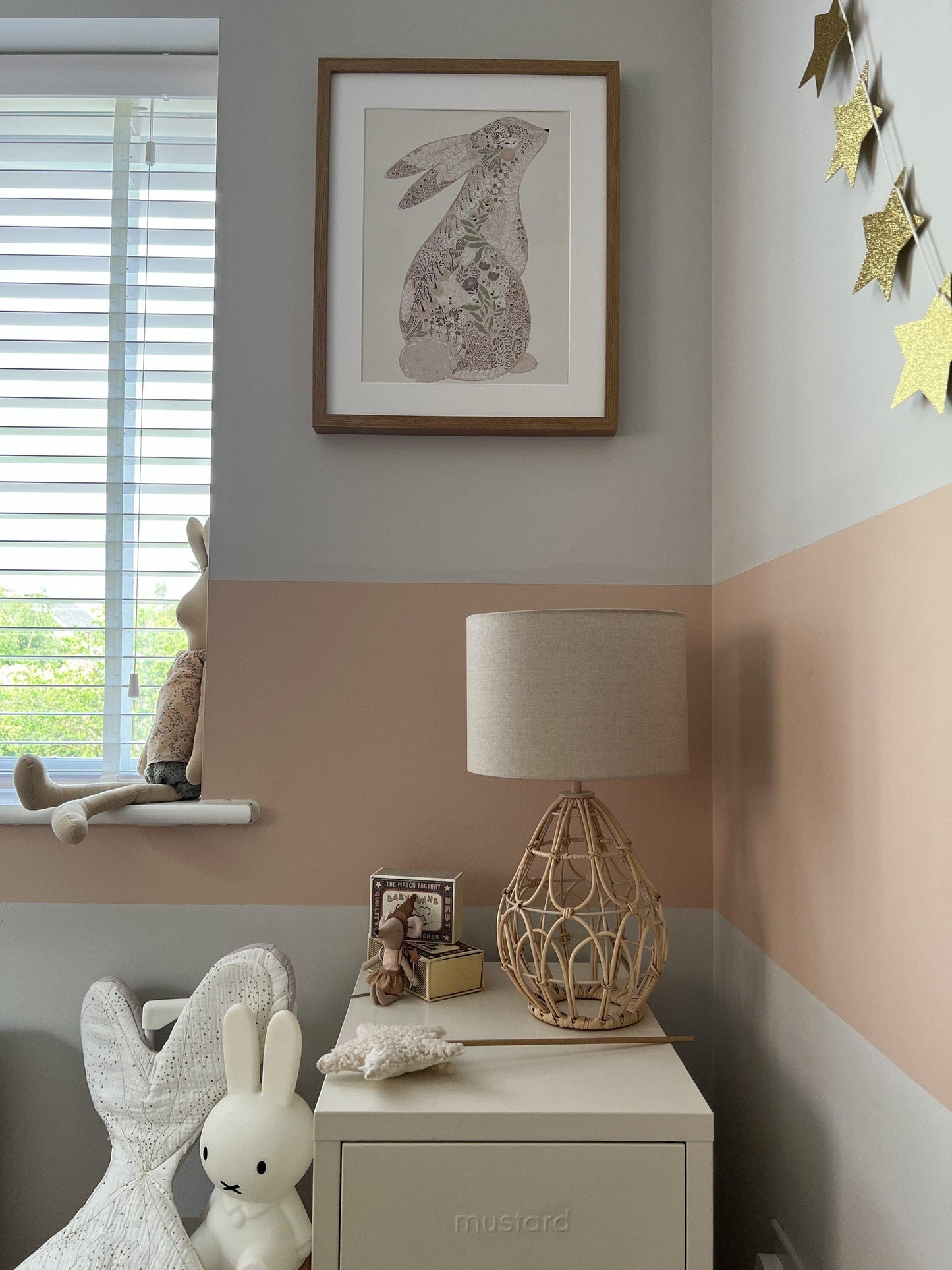 A phot of the corner of a girls bedroom, with a wide stripe of dusty pink paint halfway up the wall. A rattan based frame, maileg mice and a star wand sit on a Mustard locker in beige, with a Miffy lamp on the floor next to it. A long soft bunny sits on with window sill on the edge of the photo and our Floral bunny art print is hanging on the wall opposite the camera in a dark oak frame . while gold star bunting hangs on the wall to the right.