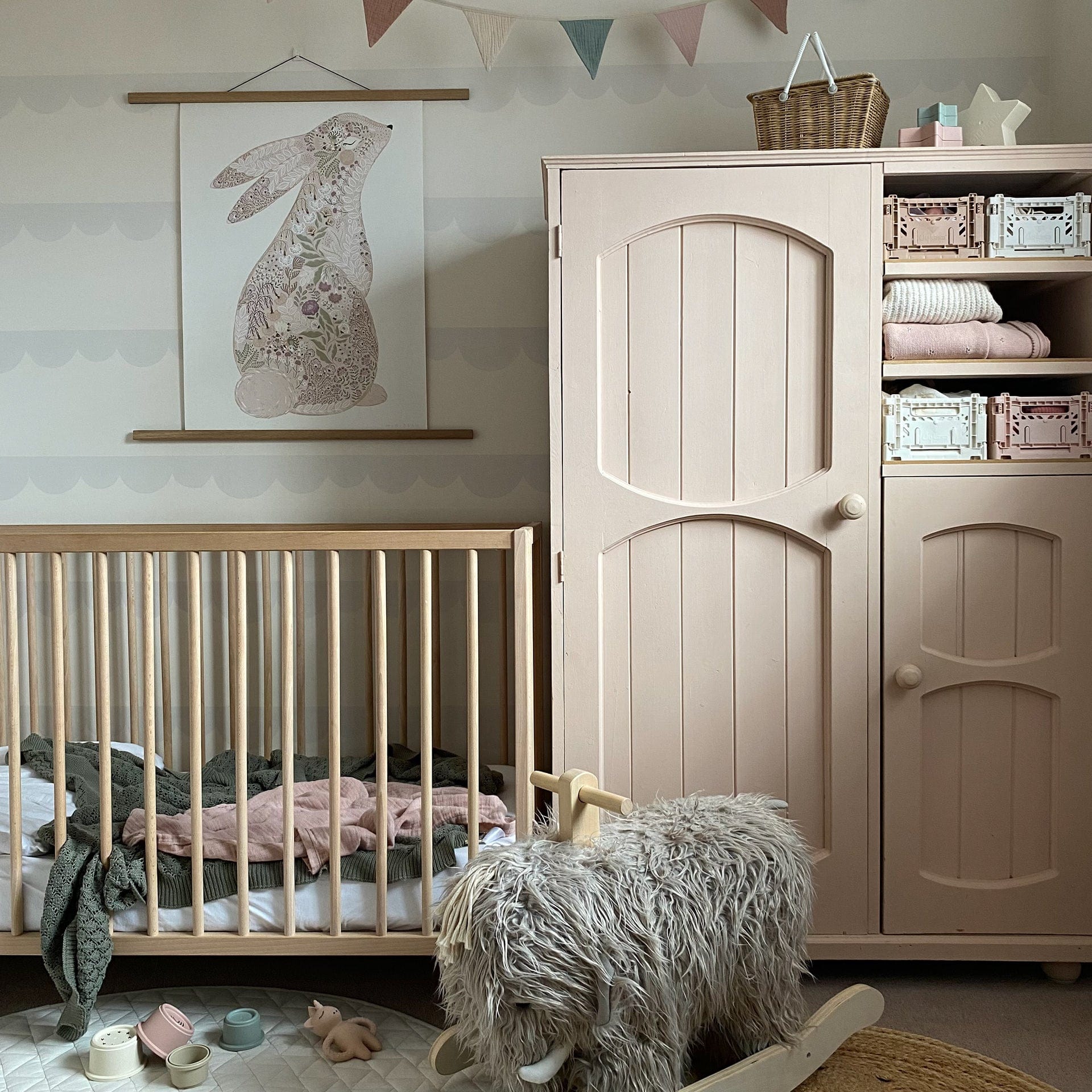 A wooden cot, with blankets strewn inside, and vintage wooden kids wardrobe, painted in a dusty pink, against a wall with scallop wallpaper, with a Kids Concept Woolly Mammoth rocker and Liewood bath toys in the foreground. On the wardrobe shelves, there are Aykasa Crates and on top of the wardrobe is an Olli Ella Piki Basket. Over the cot in a wide oak hanger is our Floral Bunny art print in pink.