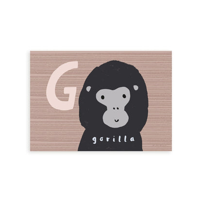 Art print in a light wood frame, leaning against a white wall, standing on parquet flooring. Our G is for Gorilla art print features a smiling cartoon gorilla from the shoulders up, on a brown wood effect background with a large G in a pale brown next to the Gorilla's head.