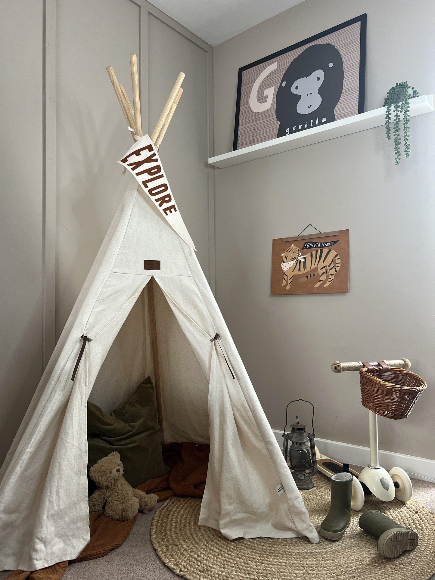 A cream Nobodinoz teepee, set up with blankets and cushions inside, with a cream triangle flag with EXPLORE in brown hanging from the top, with a black lantern, wellies, and a Banwood scooter next to it. On the wall, in an oak hanger is our forever fearless art print, and above that on a white shelf is our G is for gorilla art print and a hanging succulent.