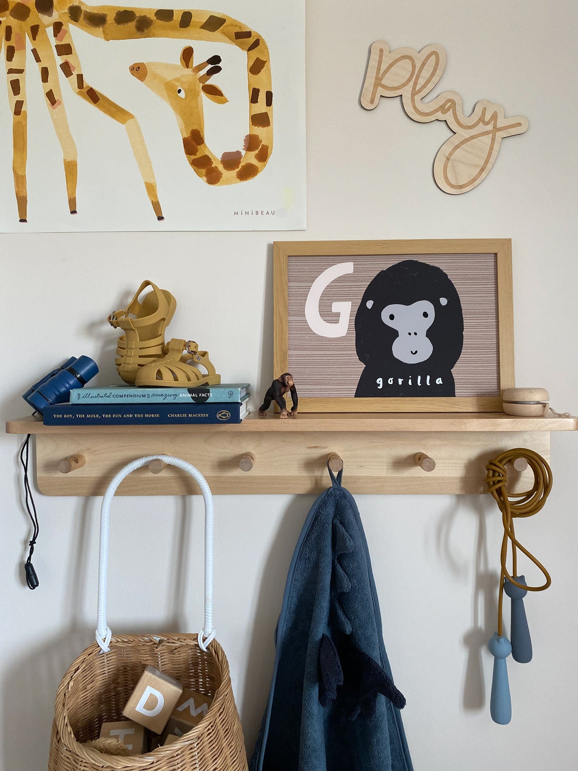A wooden shelf with wooden pegs underneath with a Liewood blue dinosaur towel, and liewood blue cat skipping rope and Olli Ella Luggy with wooden alphabet blocks in hanging, with books, liewood mustard bre sandals, a toy gorilla, a wooden yoyo and our G is for gorilla art print in an wooden frame. On the wall above is our Giraffe art print and a wooden cutout of the word PLAY.