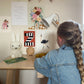 Girl sitting at her desk with a Miffy lamp on it with a selection of art prints stuck to the wall under Nobodinoz rose bunting. Featuring our boho floral vase art print, less plastic more love art print, Happy alphabet 'z' art print, party ostrich art print on the wall and the Garden art print on the desk.