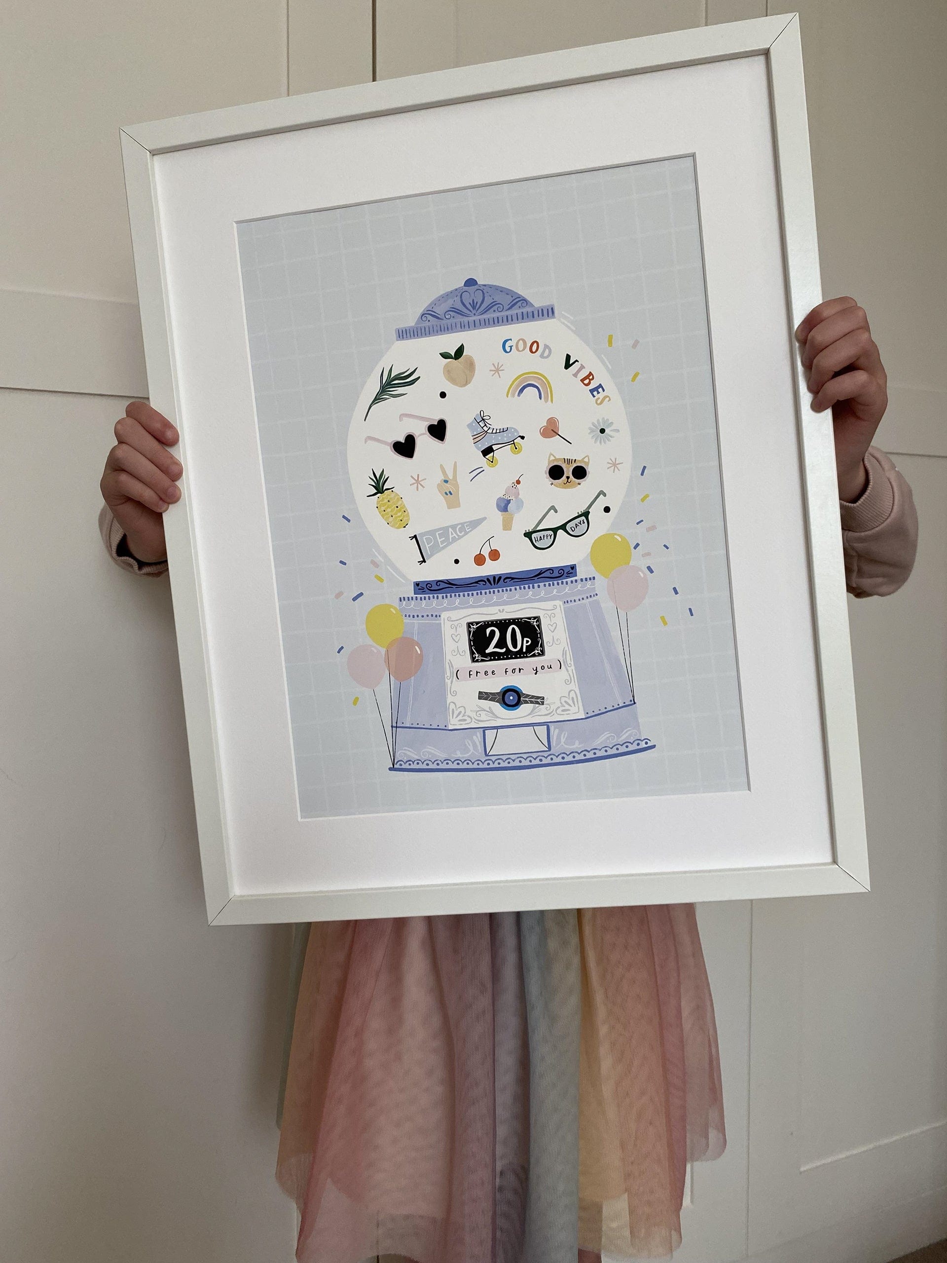 Child in a muted rainbow coloured skirt, holding up a mounted art print in a white frame. Our Good Vibes Gumball Art Print shows a vintage style gumball machine with balloons on either side on a blue background with white line check design. Inside the glass ball of the gumball machine are the words good vibes, sunglasses, roller skates, a cat in sunglasses, ice cream, a rainbow and more.