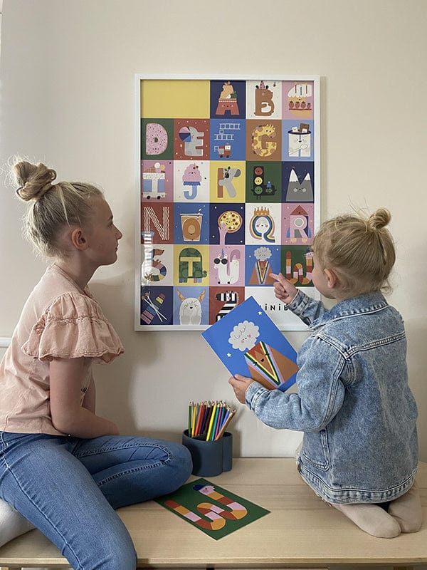 2 girls sitting looking at our Happy alphabet art print which is hung on the wall in a white frame. The younger girl is holding the Happy alphabet 'v' art print and pointing to the same image on the alphabet print. Between the girls is the happy alphabet 'w' and a liewood pot with pencils in.