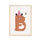Art print in a light wood frame. Our Happy Alphabet 'B' Art Print shows a brown bear with a white tummy in the shape of a B in a pink party hat with red stars on, with a bee buzzing around it's head. On a neutral background.
