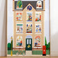 A large art print leaning against a white wall, stood on parquet flooring. Our House Art Print shows a large brick town house with 11 windows each showing a different scene, from an ironing dog to a dinosaur in the attic to the artist herself hard at work. Features a bike against a railing, a post box, traditional lamppost and hidden birds.