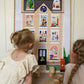 2 girls sitting on a jute ruge with alphabet blocks between them looking at our Hourt art print. Our House Art Print shows a large brick town house with 11 windows each showing a different scene, from an ironing dog to a dinosaur in the attic to the artist herself hard at work. Features a bike against a railing, a post box, traditional lamppost and hidden birds.