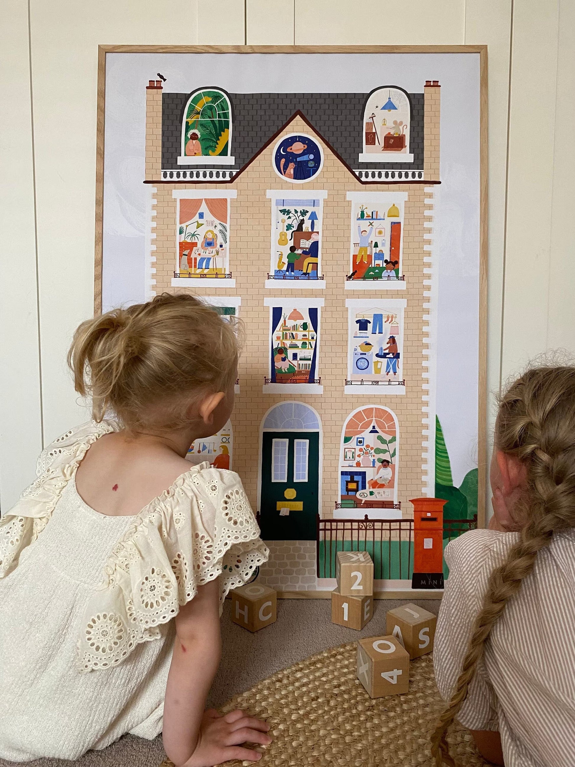 2 girls sitting on a jute ruge with alphabet blocks between them looking at our Hourt art print. Our House Art Print shows a large brick town house with 11 windows each showing a different scene, from an ironing dog to a dinosaur in the attic to the artist herself hard at work. Features a bike against a railing, a post box, traditional lamppost and hidden birds.