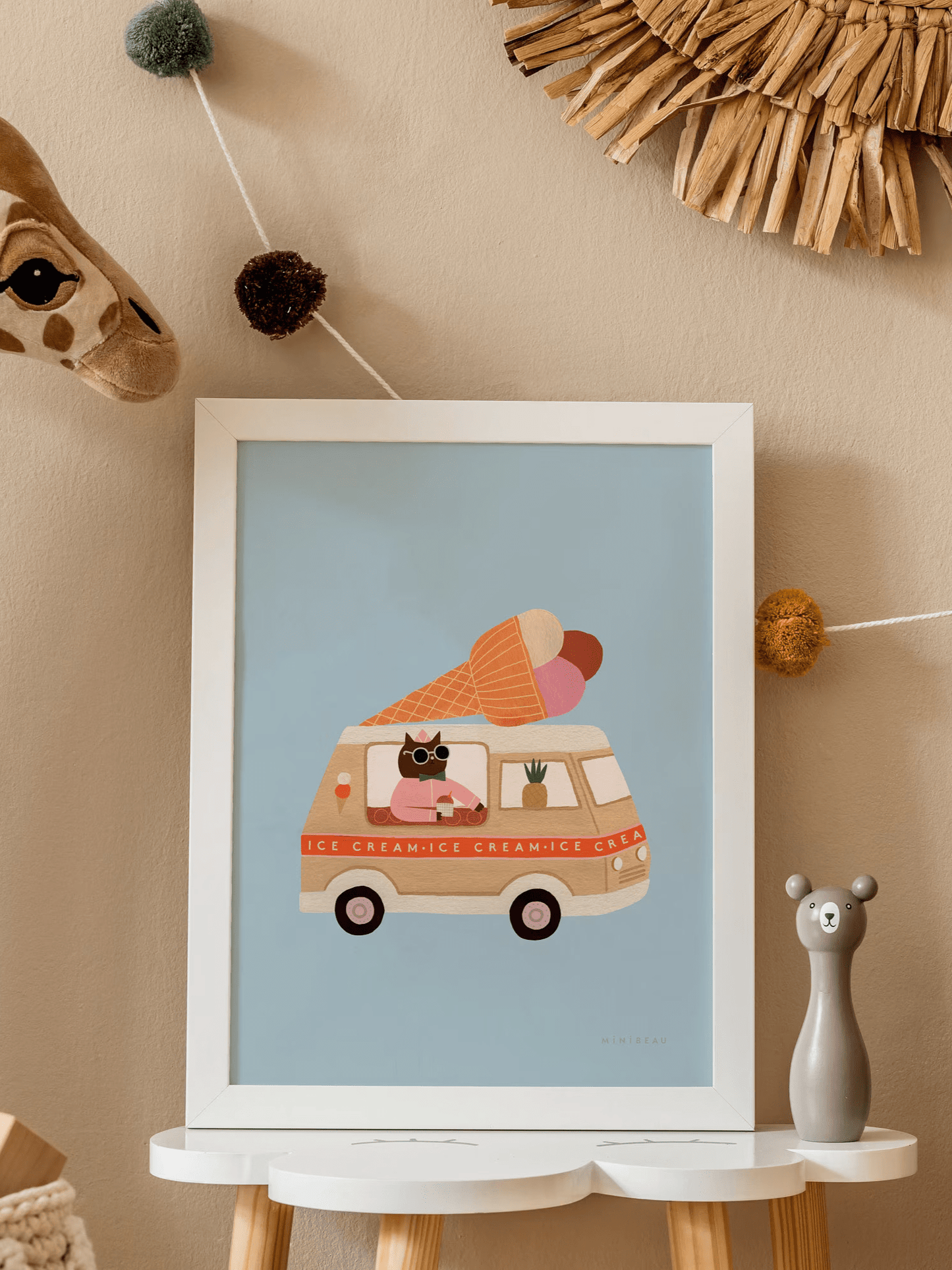 A cloud shaped side table with our Ice cream van art print on it in a white frame. With a stand up soft giraffe head in the top left and pop pom bunting in the top right. Our Ice Cream Van Art Print shows a beige ice cream van with a red stripe with the words ICE CREAM repeated along it and a large ice cream cone with 3 scoops of ice cream, chocolate vanilla and strawberry. Driven by a cool brown cat in pink and a pineapple in the front window, on a light blue background
