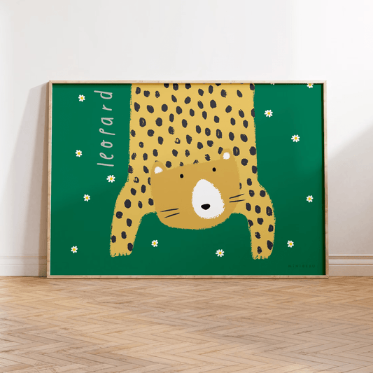 Our Leopard art print in a wooden frame leaning against a white wall, standing on parquet flooring. Hand-drawn leopard hanging down in to the picture, lifting it's head to look out at us on a green background with daisies falling.
