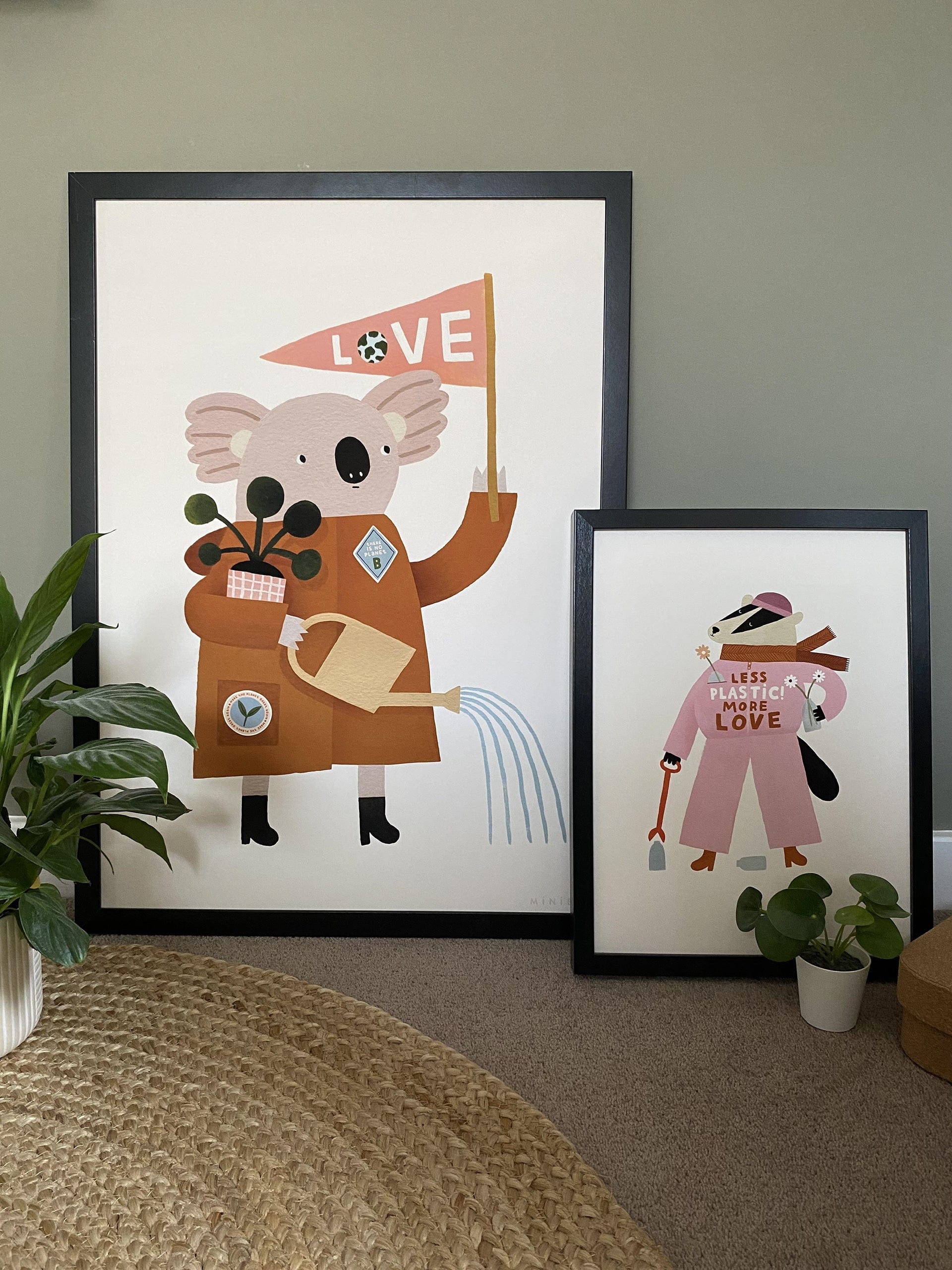 Our Love our planet art print in a large black frame, and our less plastic more love art print leaning on it in a smaller black frame, in front of a green wall, with a round jute rug and plants in the foreground.
