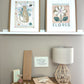 Dark wood vintage unit with a book, maileg mice, a star wand, a wooden plaque saying Autumn and a rattan based lamp, under a white shelf. On the white shelf sit 2 art prints in oak frames, our Wild, Wonderful World Leopard Art print and the Mercado de Flores art print. On the wall above the unit is our My wish for you art print in cream.