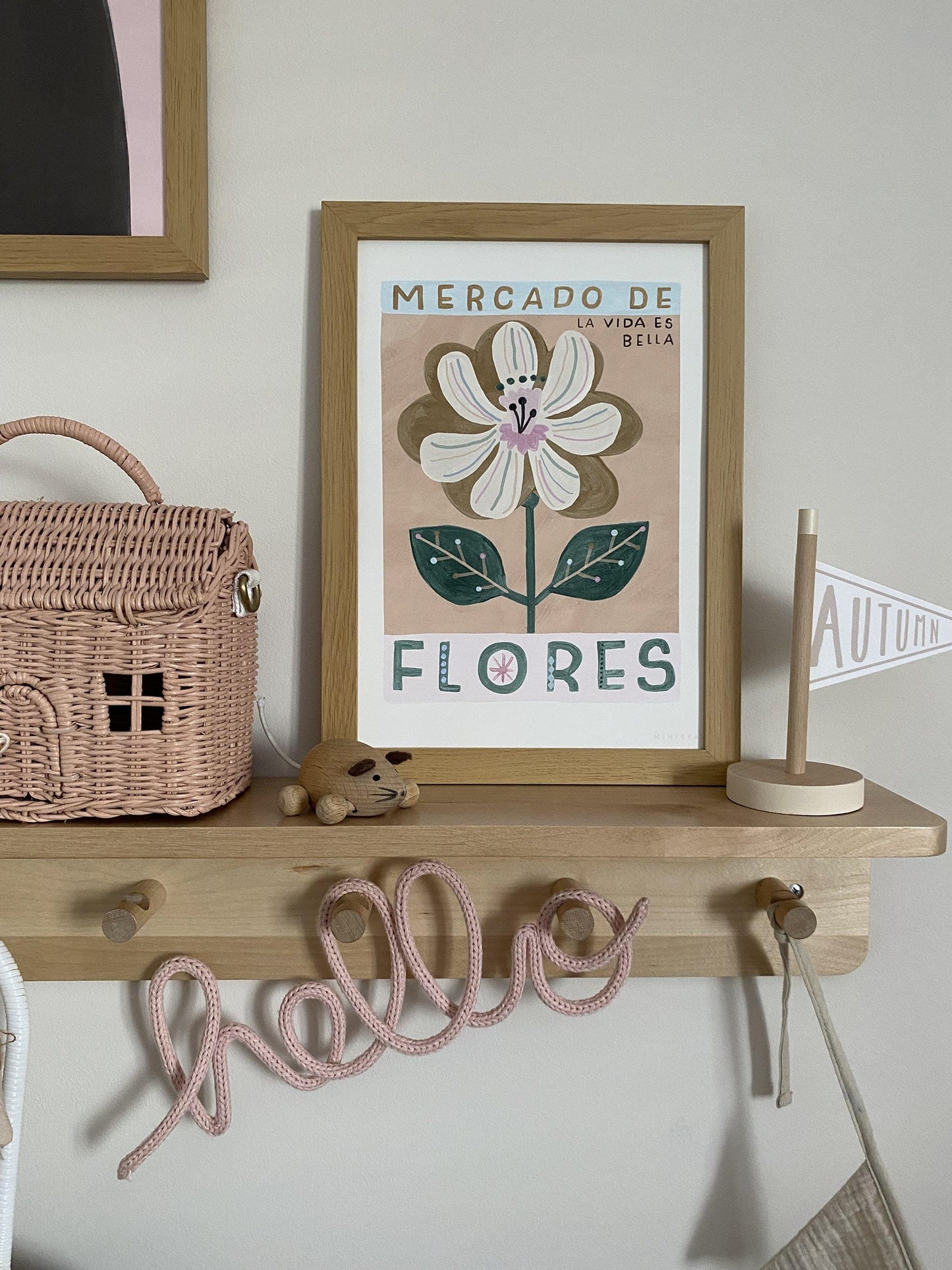 Showing one side of a wooden shelf with hanging pegs underneath with a wrapped wire word sign - HELLO, hanging on them. On the shelf is a Wooden flag saying Autumn, a wooden mouse, and Olli Ella Casa Clutch and our Mercado de flores art print in an oak frame.
