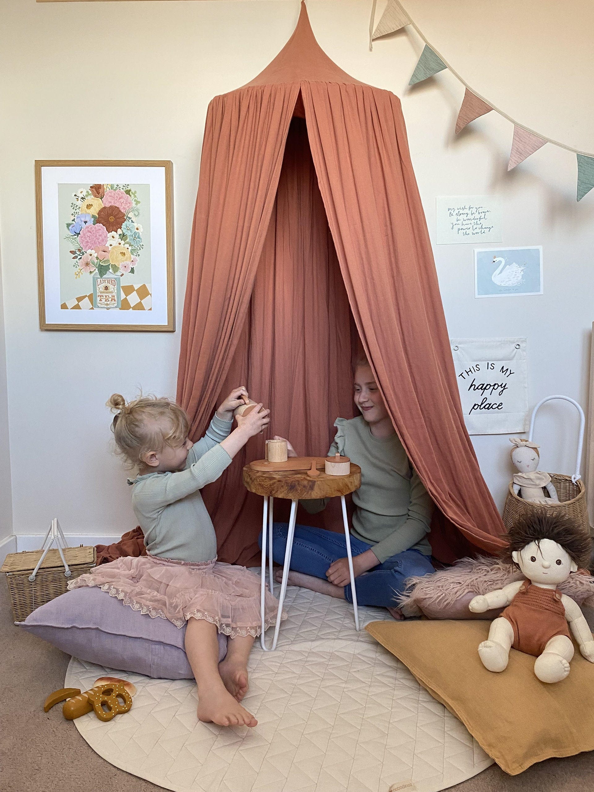 Two girls sitting on floor cushions under a rust coloured canopy, having a tea party with a wooden tea set from liewood and an Olli Ella doll in the foreground with the Boho Floral Vase art print, the wonder of you art print, and my wish for you art print on the wall behind.