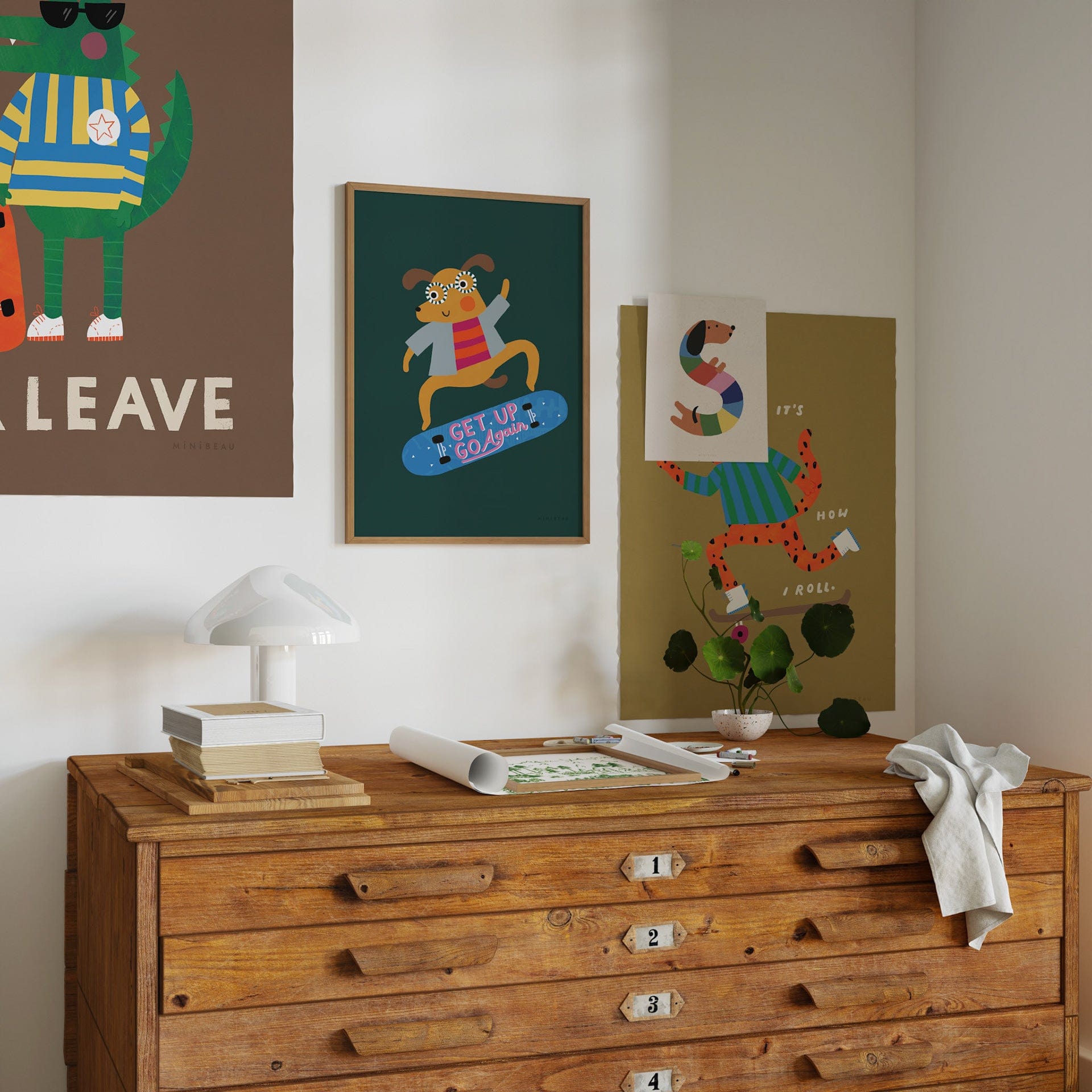 A vintage wooden chest of drawers, with a lamp and books laying on top of it and our wood art print ready to be cut to size of a light wood frame. On the wall behind hang a framed rad dog art print, it's how I roll art print, with our happy alphabet 's' art print over the cats face, and our be cool an leave art print going off the top left hand corner of the photo.