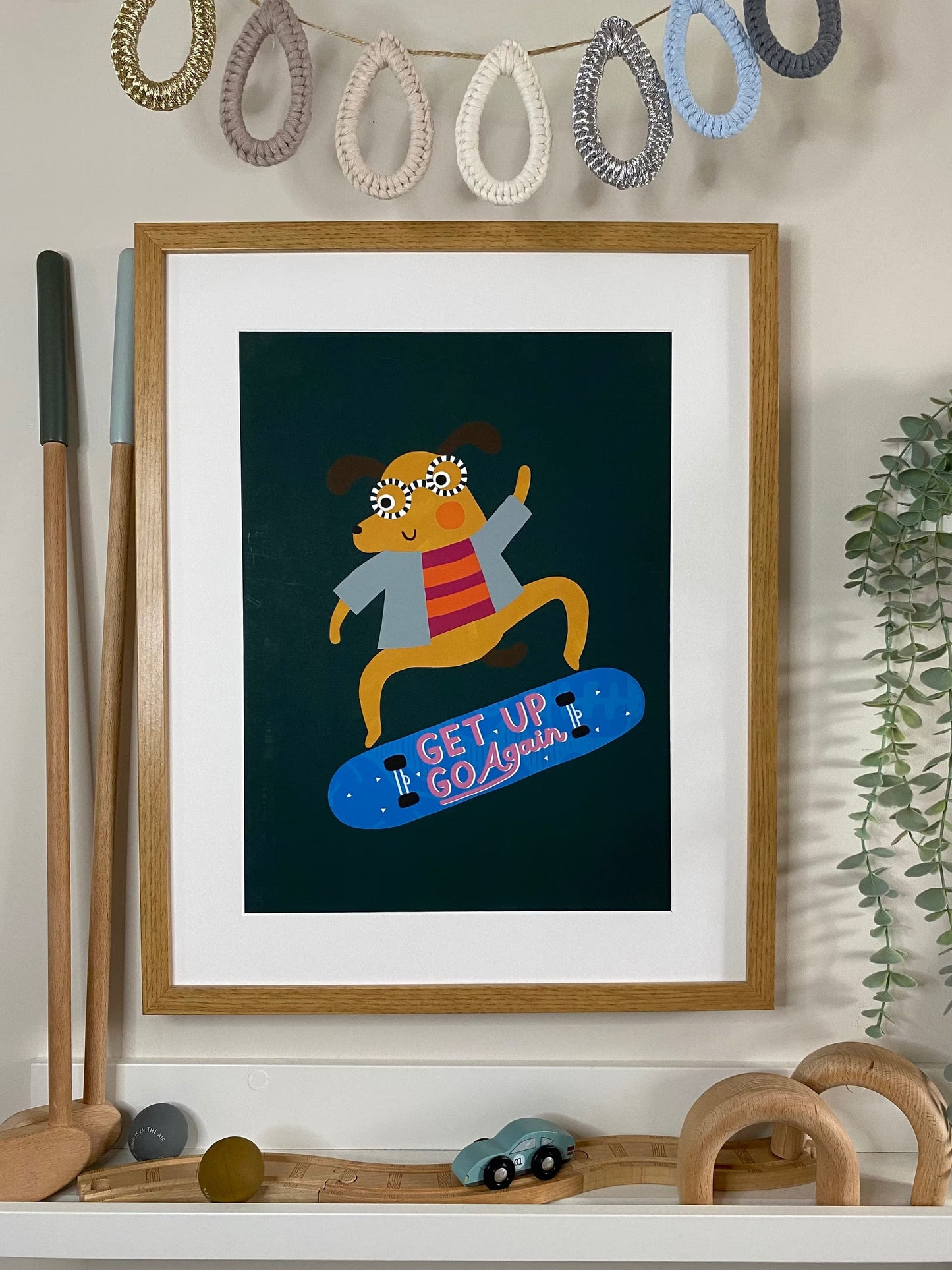 Our rad dog art print in an oak frame with a white mount low on a wall in a kids play area. with blue raindrop bunting, a vintage wooden toy race car on a wooden train track and a Liewood Mary wooden gold set. Our Rad Dog Art Print features a brown dog doing a flip on a skateboard. The dog is wearing a denim blue jacket and a bright pink and orange top while wearing black and white striped glasses on a dark green background.