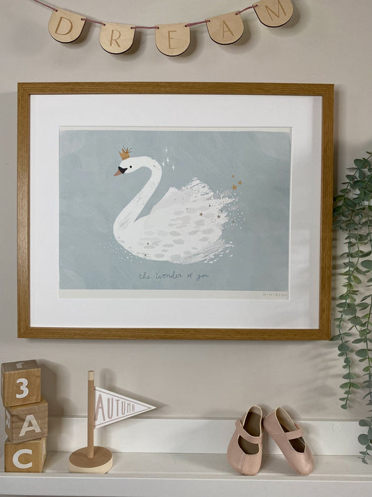 Art print in an oak frame, hung above a white shelf with wooden alphabet blocks, a wooden flag saying Autumn and some toddler shoes, with wooden bunting above the frame saying DREAM. Our The Wonder of You art print features a swan on a textured light blue background (lake). The swan is wearinga gold sparkling crown and there are stars being left behind the swan as it swims in gold and white. The wonder of you is written in a darker blue underneath the swan. The art print has a white border.