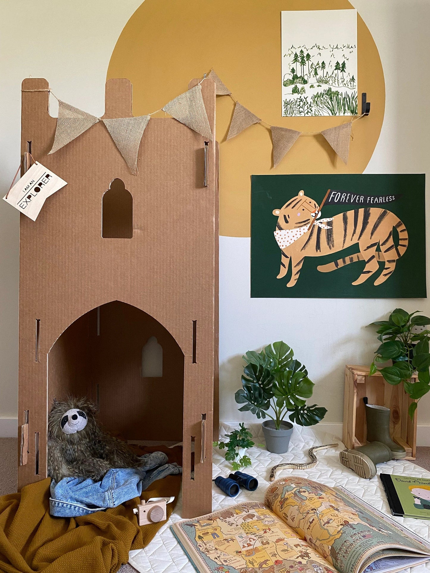 A room set up for exploration play with a Pursuit of adventure turret, a kids atlas, binoculars, plants and jute bunting with a large mustard circle painted on the wall with our woods art print and forever fearless art print in green on the wall.