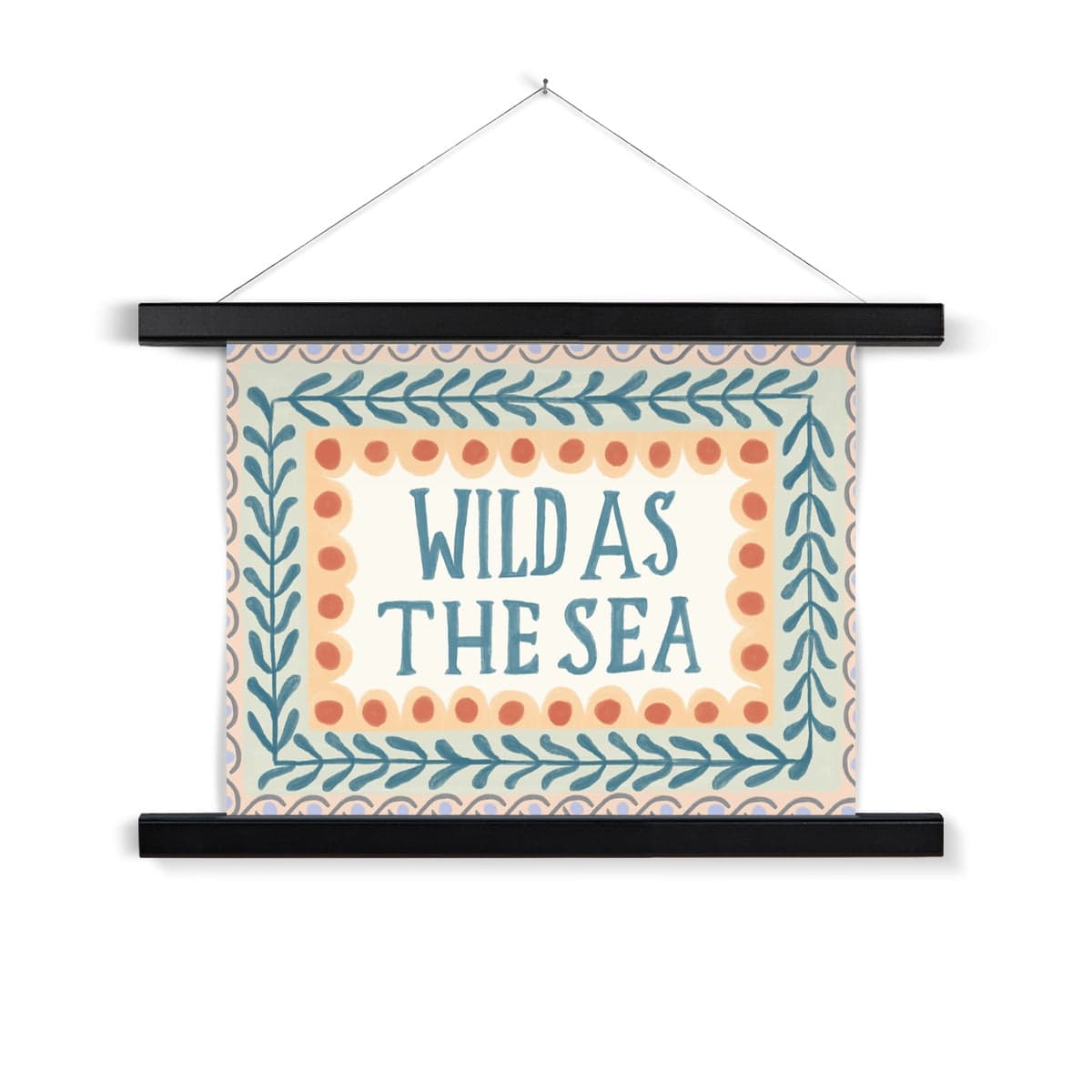 A typography print with the words WILD AS THE SEA in capital letters in watercolour deep teal in the middle on a neutral background, with 3 borders. The first being yellow scallops with deep orange dots, the second being foliage in the same colour as the typography on a pale green background and the outer being a sage wave pattern with lilac dots on a peach background, hanging from a nail in a black wood hanger