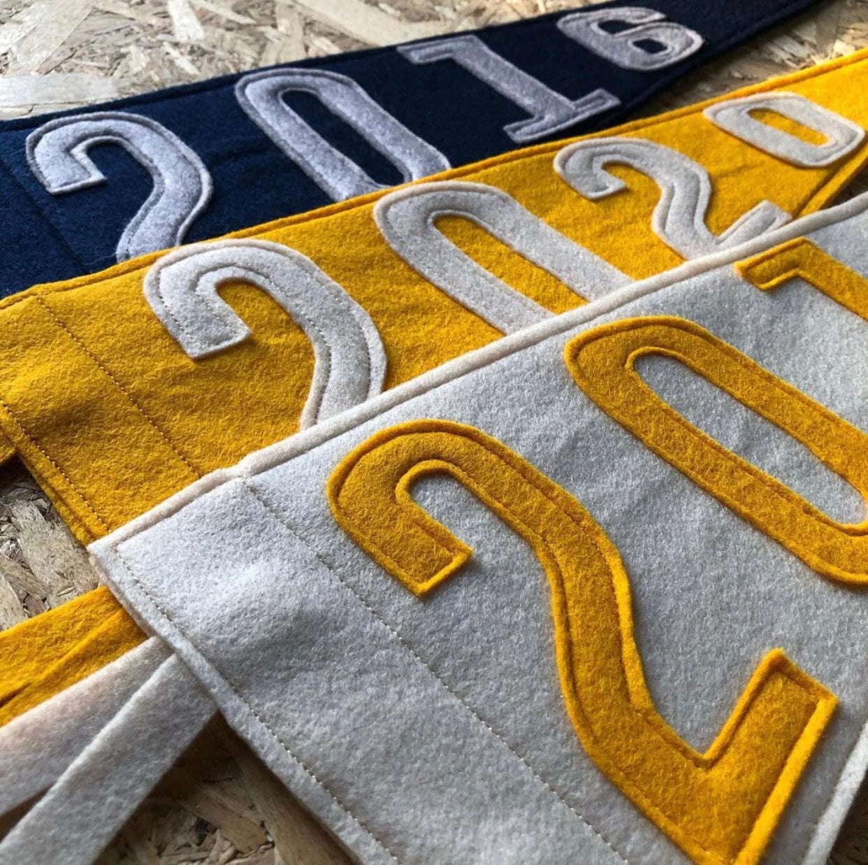 3 pennants laying on wood, over-lapping, 1 dark blue with grey numbers, one yellow with off white numbers and one off white with yellow numbers.