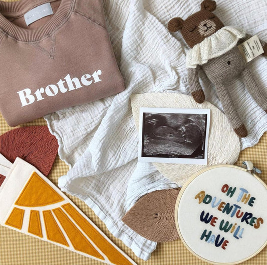 Flat lay showing the sunshine pennant - narrow off-white triangle with 2 ties, with 5 sunbeams coming from a yellow quarter circle on the top left of the triangle - with a baby scan, brother sweatshirt, teddy, and muslins