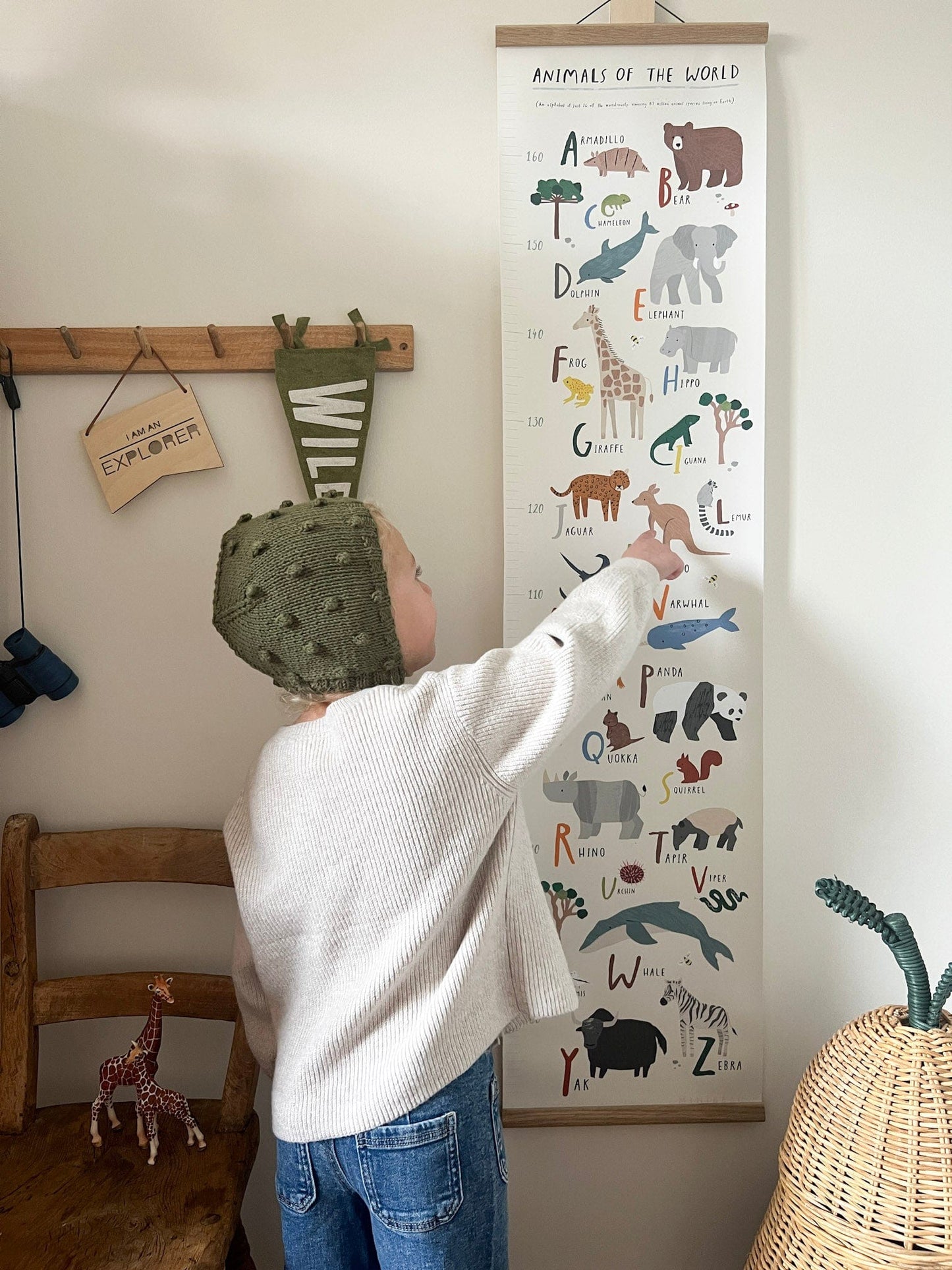 Child pointing at the kangaroo on the Animals of the world height chart in a solid oak hanger on a wall next to wooden vintage coat hooks with a wild flag hanging from it, next to a vintage wooden kids chair with a denim jacket hanging over it and a ferm living pear basket.
