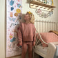 Blonde girl standing in front of the folk carnival height chart and looking back at it. Vintage metal framed bed in the background and a wooden shelf with peg hooks with a miffy lamp on it and raindrop bunting