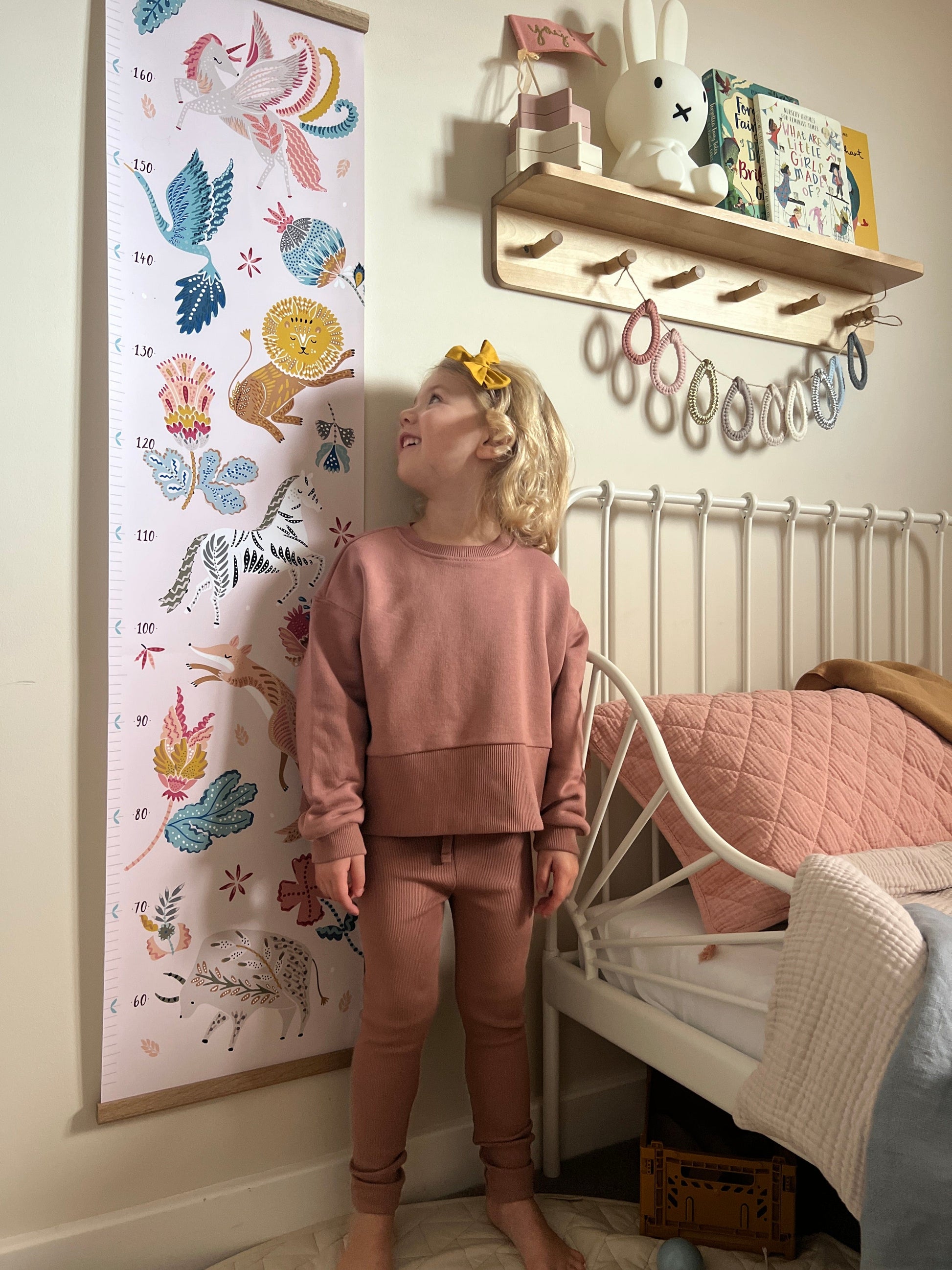 Blonde girl standing in front of the folk carnival height chart and looking back at it. Vintage metal framed bed in the background and a wooden shelf with peg hooks with a miffy lamp on it and raindrop bunting