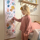 Blonde girl pointing at the zebra on the folk carnival height chart. Vintage metal framed bed in the background and a wooden shelf with peg hooks with a miffy lamp on it and raindrop bunting