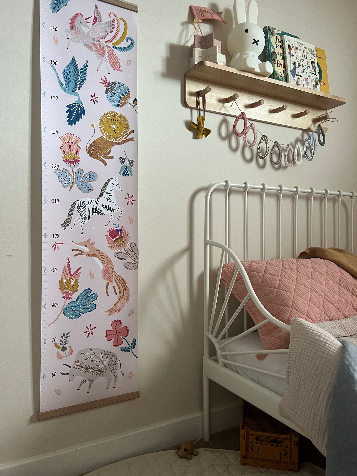Our folk carnival height chart hanging on a neutral wall. Vintage metal framed bed in the background and a wooden shelf with peg hooks with a miffy lamp on it and raindrop bunting.