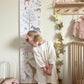 Girl in white dress with a pink bow in her hair, standing in front of height chart looking back at the animals on it, next to a cot and a shelf with hooks under it. Gold star garland is hanging down