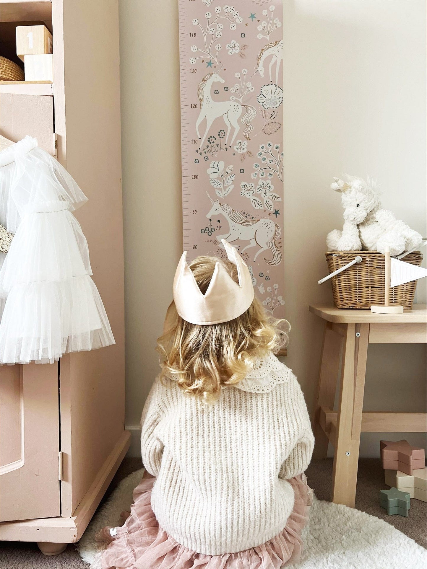 Girl in a fabric crown wearing a pink skirt and cream knit jumper, crouching in front of the unicorn height chart which is hanging between a pink vintage wardrobe and a wooden bench with a olli ella piki basket on it.