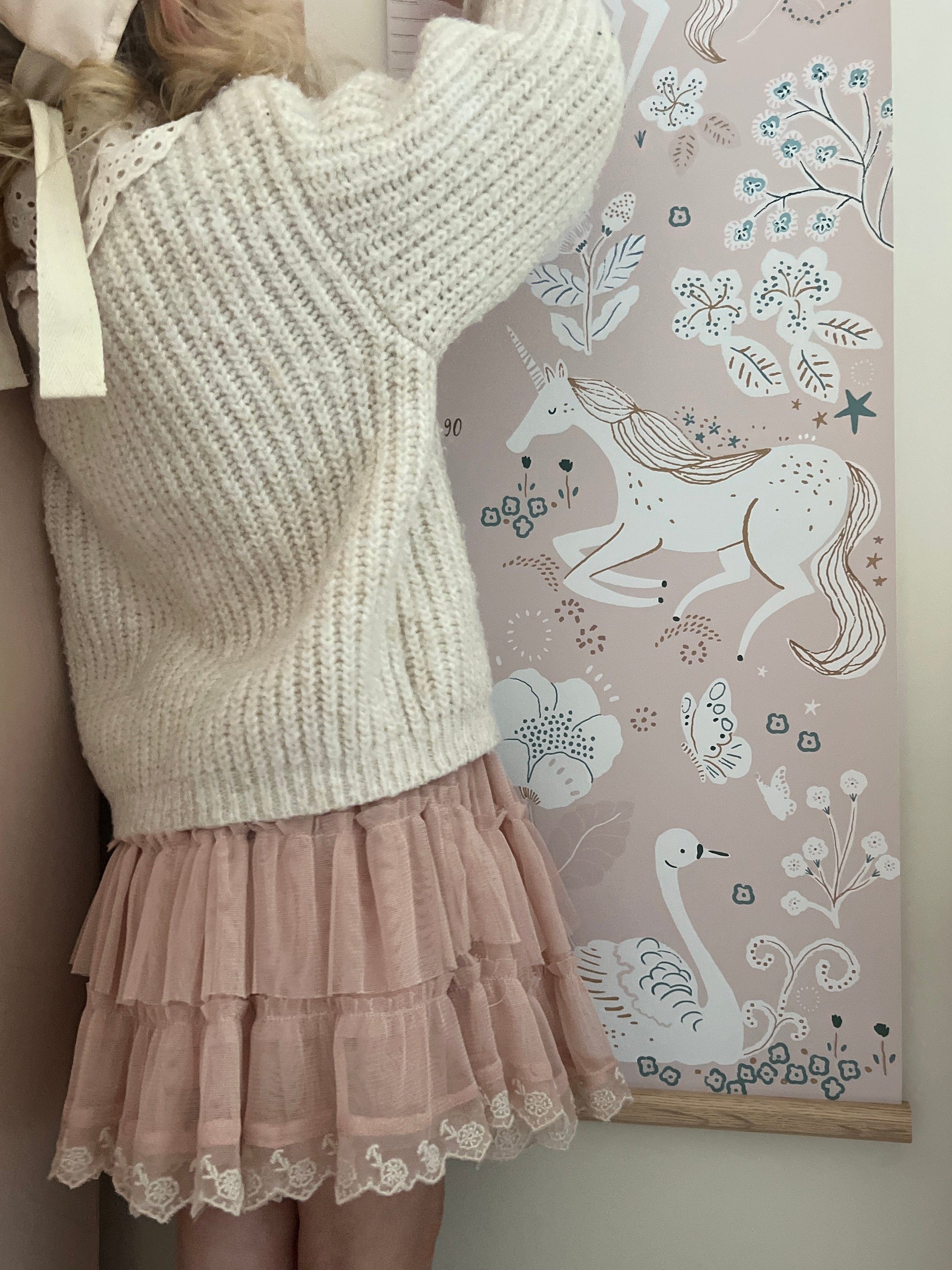 ±Girl in a pink skirt and cream knit jumper standing in front of the unicorn height chart pointing at details. Showing the oak hanger at the base of the height chart